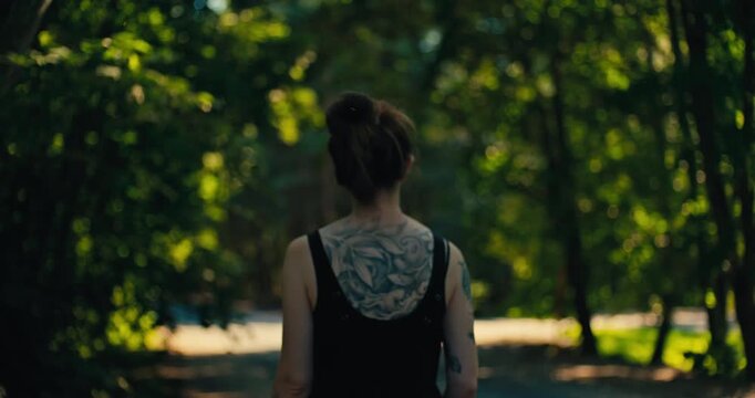Rear view of a woman walking through a sunlit woodland. Warm natural light and lush green trees create a peaceful forest setting while a detailed back tattoo is visible.