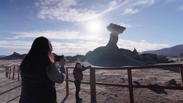 Mother and her young daughter standing at a wooden fence viewpoint next to the famous El Hongo rock formation in Ischigualasto park. Slow motion shot on sunny afternoon.