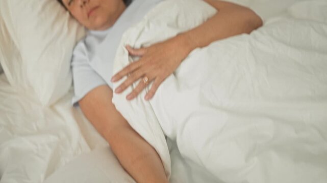 Woman sleeping in bed holding remote with hand on white duvet beside pillow and wooden headboard in a building bedroom; calm rest.