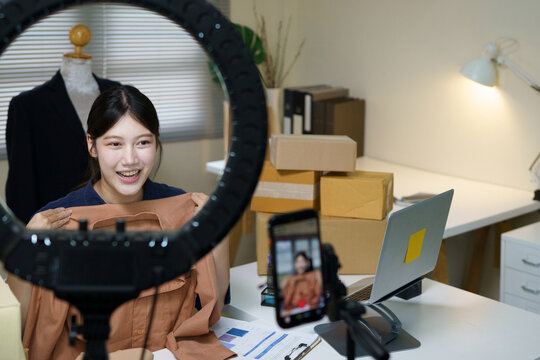 Asian woman live streaming fashion from a cozy home studio, promoting her small e commerce boutique on social media using smartphone and ring light to showcase clothes and connect with customers