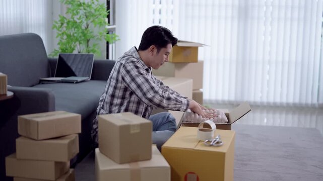 Young Asian man packing cardboard boxes with packing tape in bright apartment living room during relocation day