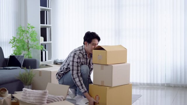 Young Asian man kneeling on floor organizing stacked cardboard boxes in bright modern apartment during move in day