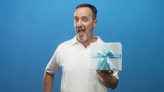 Man holding white gift box tied with blue ribbon in his hand while smiling and presenting it in studio; celebration joy.