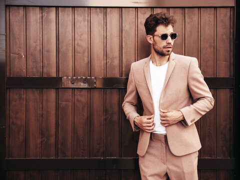 Handsome confident stylish hipster lambersexual model.Sexy modern man dressed in elegant beige suit. Fashion male posing in the street near wooden fence wall in city at sunset. In sunglasses