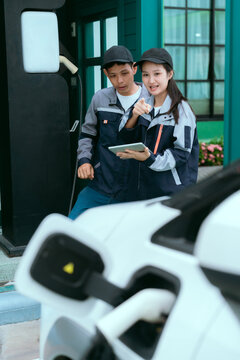 Two Asian technicians in uniform using a digital tablet to inspect and discuss the EV charging station setup.