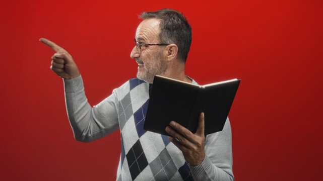 Man pointing finger with open mouth while holding book, wearing argyle sweater and glasses in studio; surprise curiosity discovery.