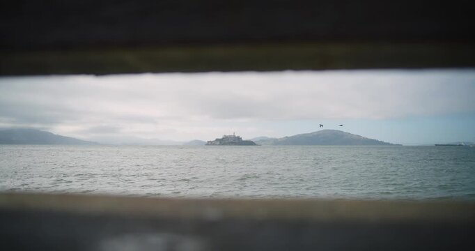 4K cinematic view of Alcatraz Island from the San Francisco waterfront near Pier 39. The historic prison island sits across the bay with surrounding water and birds in flight.