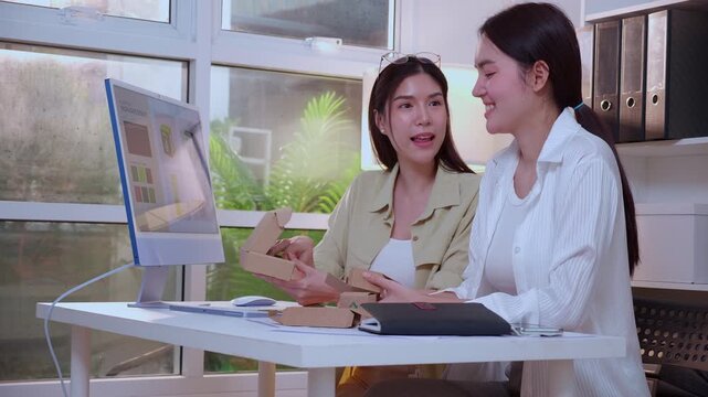 Two young asian woman designer working on packaging design with ai software with desktop computer, business team discussion of eco friendly cardboard box product development at modern home office.
