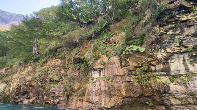 Wide panoramic view of river canyon with sheer layered cliff rising from teal water, damp seepage staining rock face, soft surface ripples, and dense deciduous growth across upper slopes