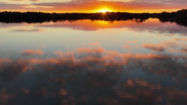 Aerial video gliding over a calm bay as the colours of the sunset sky are reflected across the glassy water, creating a peaceful coastal scene with soft light and tranquil evening conditions.