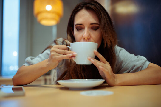 Peaceful Woman Sipping Coffee From White Cup At Cozy Cafe. Calm customer enjoys hot morning beverage sitting alone inside serene local coffee shop