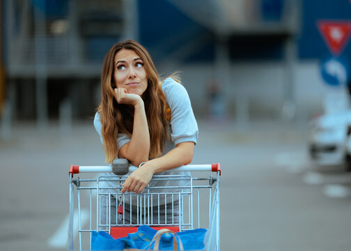 Thoughtful Woman Standing By Shopping Cart In Supermarket Parking Lot. Confused female customer looks away trying to remember what she forgot to buy
