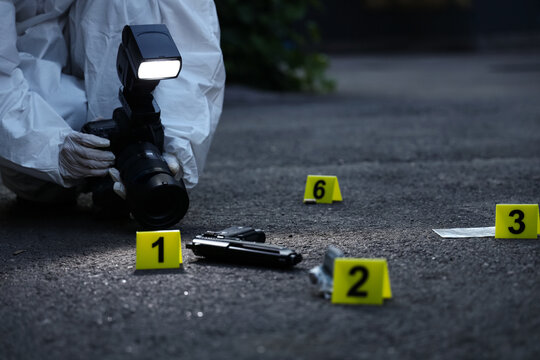 Forensic investigator in protective suit photographing crime scene evidence with Numbered markers and a handgun, Numbered yellow markers indicate items including a handgun, a bullet shell casing