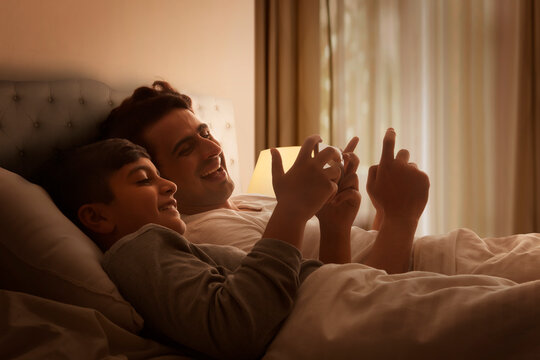 Happy little boy and his father using cell phone in bedroom