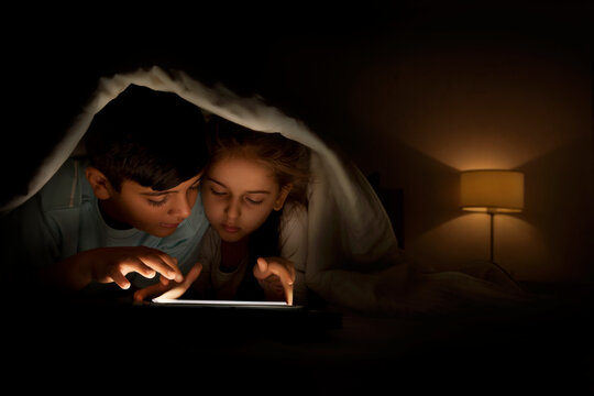 Boy and girl sharing digital tablet lying in bed under the covers