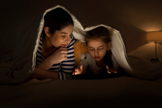 Mother and daughter using digital tablet together lying in bed under the covers