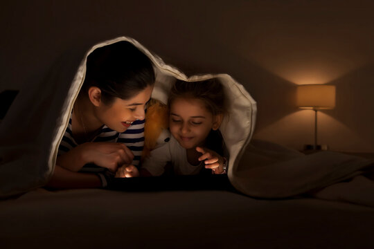 Smiling mother and daughter using digital tablet together lying in bed under the blanket