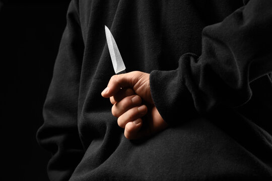 Person in black hoodie holding a sharp knife behind their back, betrayal, danger, and hidden threat