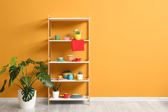 Wooden shelving unit with colorful kitchenware and a monstera plant against a bright orange wall, home decor and interior design