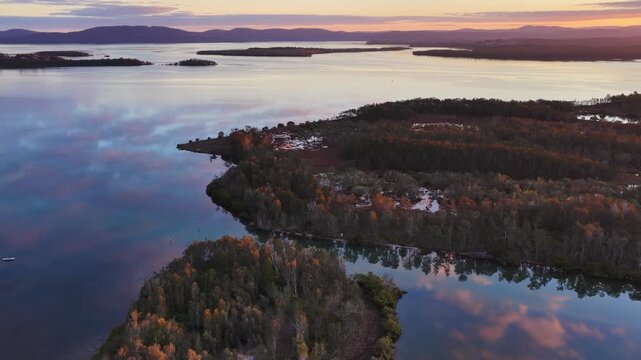 Aerial video gliding over a calm bay as the colours of the sunset sky are reflected across the glassy water, creating a peaceful coastal scene with soft light and tranquil evening conditions.