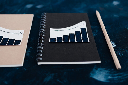 black and kraft notebooks with rising bar graph stickers and pencil on dark blue surface