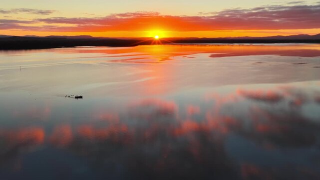 Aerial video gliding over a calm bay as the colours of the sunset sky are reflected across the glassy water, creating a peaceful coastal scene with soft light and tranquil evening conditions.