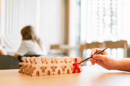 Hand using paintbrush to paint one wooden figure red among a group. Ideal for talent selection, employee development, HR recruitment, and creating uniqueness commercial business concepts.