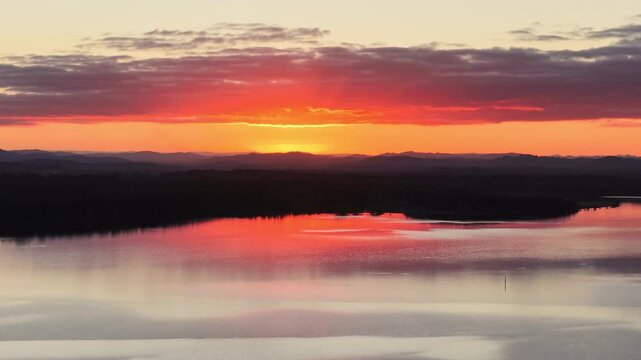 Aerial video gliding over a calm bay as the colours of the sunset sky are reflected across the glassy water, creating a peaceful coastal scene with soft light and tranquil evening conditions.