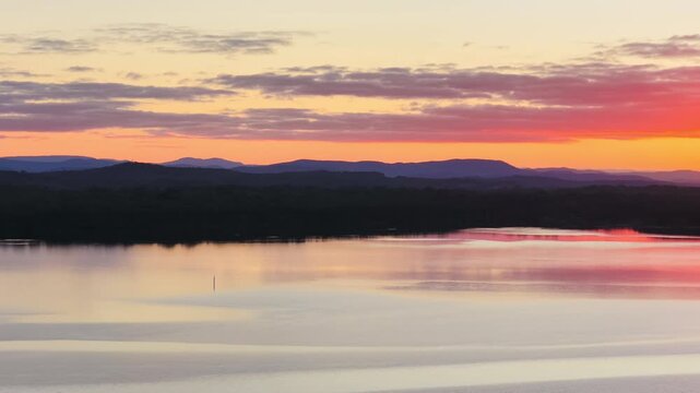 Aerial video gliding over a calm bay as the colours of the sunset sky are reflected across the glassy water, creating a peaceful coastal scene with soft light and tranquil evening conditions.