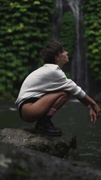 Young woman in a sweatshirt crouching by a waterfall