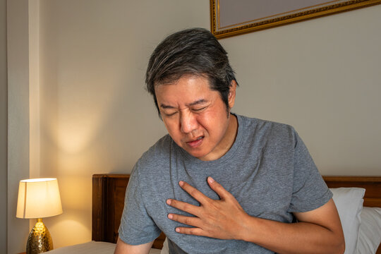 Man sitting on bed with hand on chest, depicting sharp chest pain from heart and cardiovascular disease for medical treatment concept.