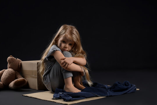 Homeless little girl sitting on piece of cardboard against dark background