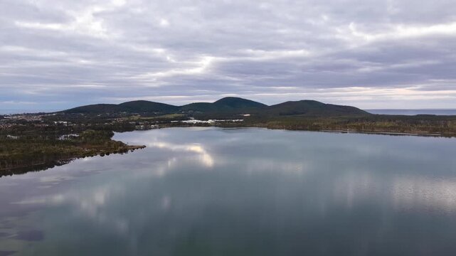 Aerial video gliding over a calm bay as the colours of the sunset sky are reflected across the glassy water, creating a peaceful coastal scene with soft light and tranquil evening conditions.