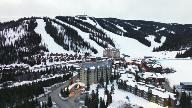 Dolly in tilting up aerial drone wide shot of a winter landscape of a mountain resort in Montana surrounded by pine forests, homes, and snow-covered terrain at dusk.