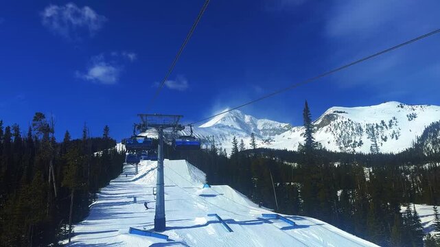 Handheld winter footage from a ski lift overlooking a Montana mountain landscape with Lone Peak in the distance, snowy trails below, pine forests, and lift chairs passing on a sunny March day.