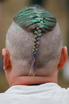 Back view of a person with a shaved head. Single green braided hair strand extends from the crown. I