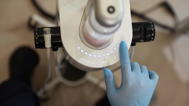 Overhead clinical close-up of dental microscope head with gloved operator pressing illuminated control on numbered dial, blue indicators, precision equipment and sterile treatment room