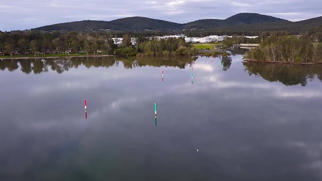 Aerial video gliding over a calm bay as the colours of the sunset sky are reflected across the glassy water, creating a peaceful coastal scene with soft light and tranquil evening conditions.