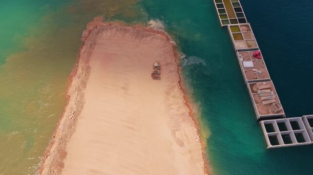 Aerial top-down view of a bulldozer pushing sand and earth into the turquoise sea, reclaiming land for a coastal construction project near floating structures on a sunny day