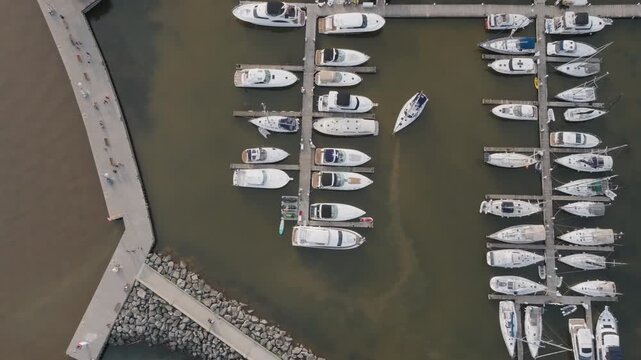 A high top-down perspective captures boats docked in neat rows along marina piers in Oakville. One white sailboat maneuvers slowly through the narrow water channel.