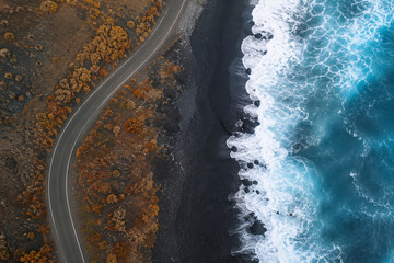 Obraz premium Aerial perspective of a winding road along a volcanic black sand beach with crashing blue ocean waves.