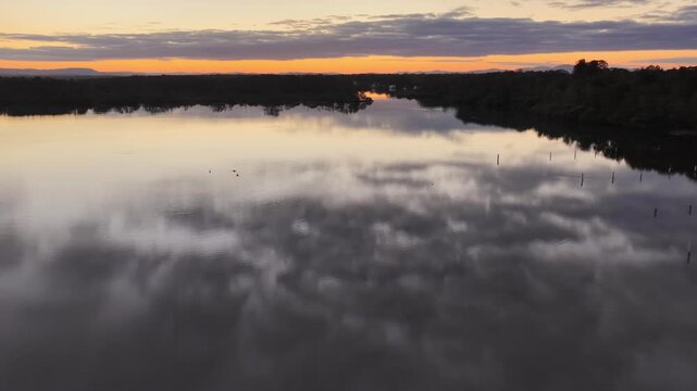 Aerial video gliding over a calm bay as the colours of the sunset sky are reflected across the glassy water, creating a peaceful coastal scene with soft light and tranquil evening conditions.