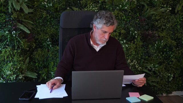 Focused senior man working on laptop computer and checking printed papers at office desk. Concept for data entry, accounting, solo worker, report review, and concentration.
