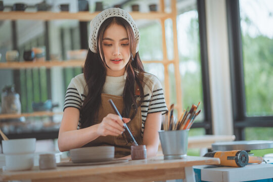 Happy Asian female artist using smartphone to find references and ideas for painting ceramic pottery design in creative studio, woman painting custom artwork for client project