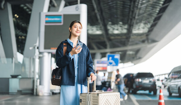 Young adult asian woman with travel luggage using smartphone app call taxi service transfer at international airport shuttle bus terminal