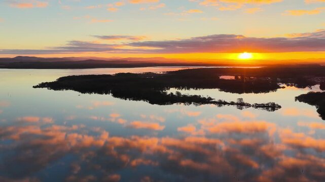 Aerial video gliding over a calm bay as the colours of the sunset sky are reflected across the glassy water, creating a peaceful coastal scene with soft light and tranquil evening conditions