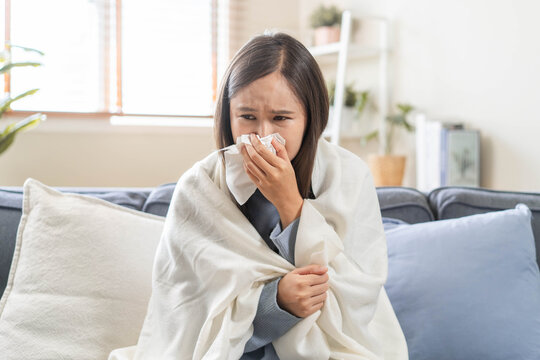 Sick Asian woman blowing her nose with tissue on sofa, wrapped in blanket, suffering from high fever, seasonal flu, influenza virus infection, and cold symptoms at home