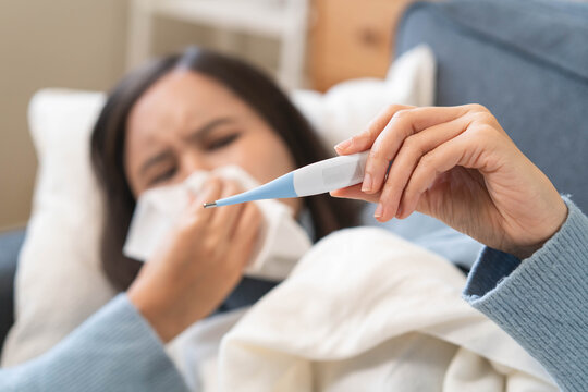 Sick person resting in bed checking temperature with thermometer, suffering from high fever, seasonal flu, influenza virus infection, and common cold symptoms at home