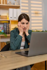 Fototapeta premium Young woman on home table reads bad news on laptop and feels stress during remote work trouble at home. Adult girl on home table reacts with disappointment and despair after seeing bad online result.