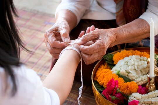 Thai culture ritual, hand tie blessing, elder hands, white string, flower tray, spiritual way, ceremonial care and quiet respect in warm home setting